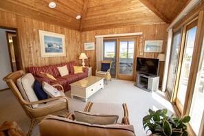 A wooden-floored living room with a sofa, chairs, a TV, and a balcony with glass doors.
