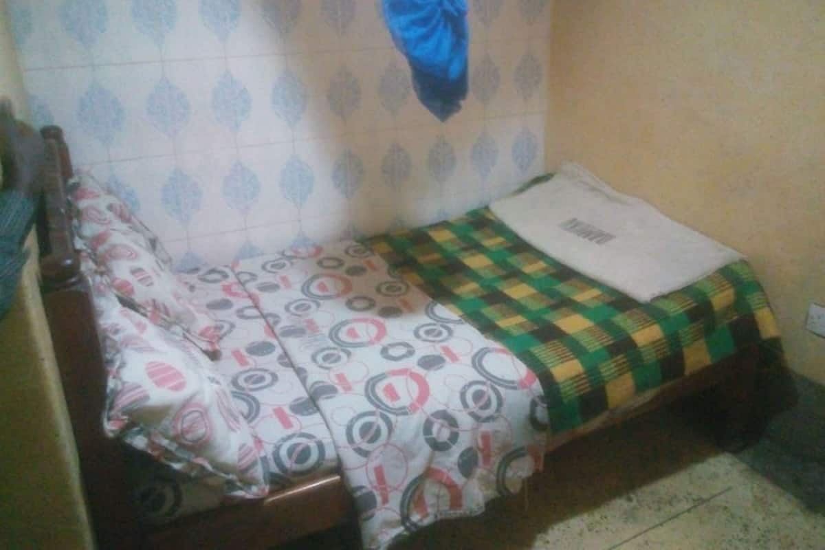 A small, single-bed room with a patterned bedspread, a plain pillow, and a blue garment hanging on the wall.