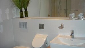 Ein Badezimmer mit Waschbecken, Toilette und Dusche. Auf dem Ablagetisch stehen zwei grüne Flaschen.