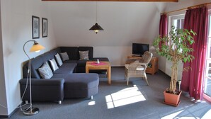 Ein Wohnzimmer mit einem Ecksofa, einem kleinen Tisch, einem Sessel, einer Stehlampe, einem Fernseher und einer Topfpflanze.