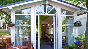 Un restaurant à la façade blanche et aux grandes fenêtres, doté d’une terrasse et de jardinières suspendues.