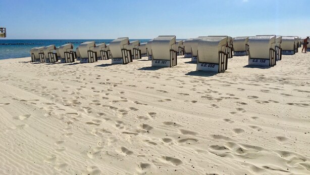 De nombreuses cabanes de plage, chacune portant un numéro, sont alignées sur une plage de sable où l’on peut voir des empreintes de pas.