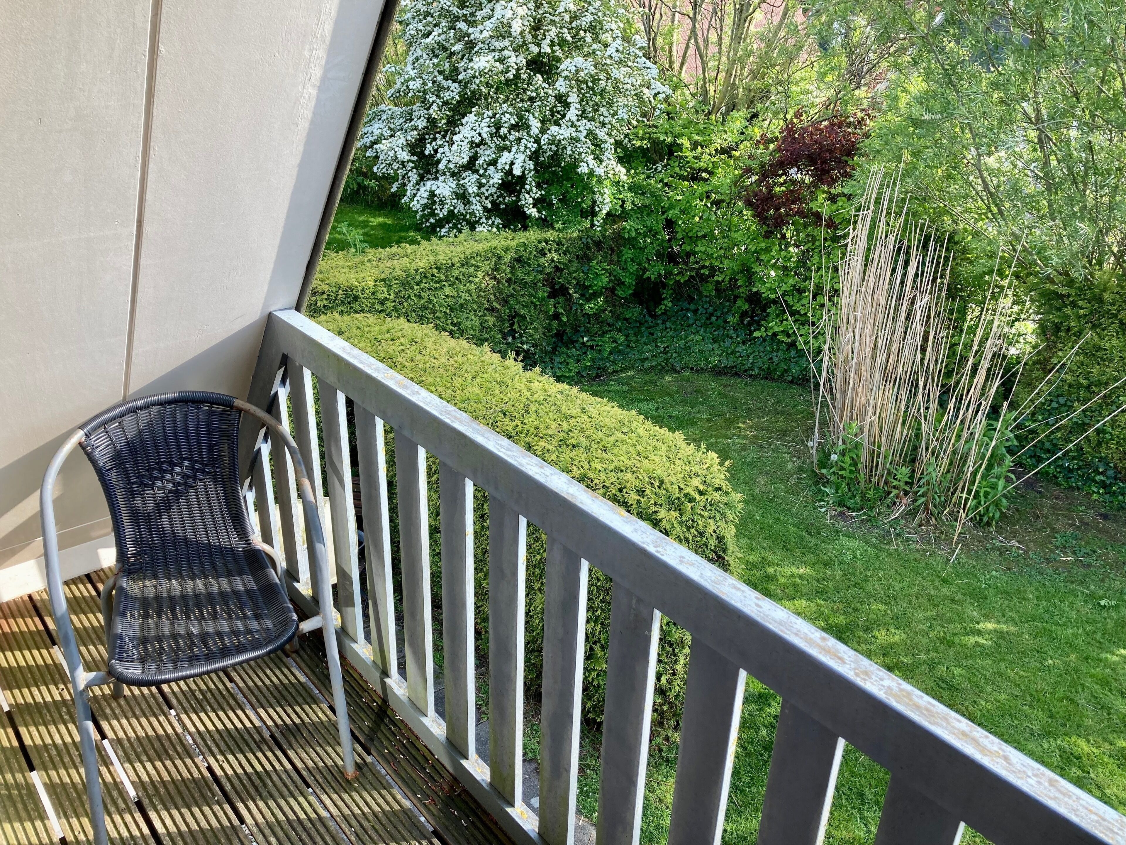 A balcony with a wicker chair, a metal railing, and a view of a well-maintained garden with various plants and a hedge.