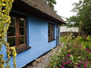Une maison traditionnelle aux murs bleus et au toit de chaume, entourée d’un jardin où poussent différentes plantes et fleurs.