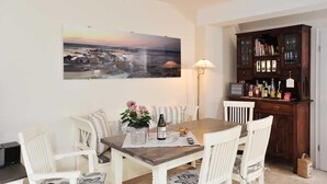 A dining area with a wooden table, white chairs, a floral centerpiece, and a large landscape painting on the wall. - Meerwievke (Sylt)