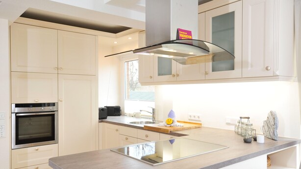 A modern kitchen with white cabinetry, a built-in oven, a stainless steel range hood, and a central island with a sink. - Meerwievke (Sylt)