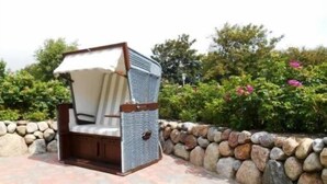A patio chair with a cushioned seat and backrest, positioned on a stone-tiled patio surrounded by a stone wall and greenery. - Meerwievke (Sylt)