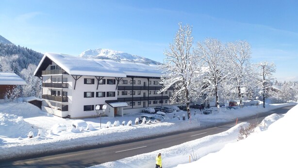 A multi-story hotel with balconies, a parking area, and a road in a snowy landscape with mountains in the background. - Ehlershus (Reit im Winkl)