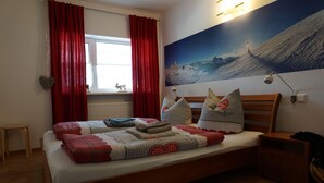 A bedroom with a wooden bed, red curtains, a mountain landscape painting, and a small table. - Ehlershus (Reit im Winkl)