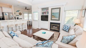 A spacious living room with a sofa set, a coffee table, and a fireplace. There is a kitchen area with a refrigerator and a dining table. - Oceanside, Community Pools, Community Tennis, Seasonal Community Trolley, M418, Mermaids Cove (Corolla)