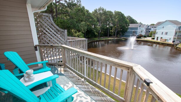 A wooden deck with turquoise chairs and a small table, overlooking a calm water body and a row of buildings. - Oceanside, Community Pools, Community Tennis, Seasonal Community Trolley, M418, Mermaids Cove (Corolla)