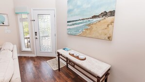 A room with a wooden floor, a bench with bowls, a painting of a beach, and a door with a window. - Oceanside, Community Pools, Community Tennis, Seasonal Community Trolley, M418, Mermaids Cove (Corolla)