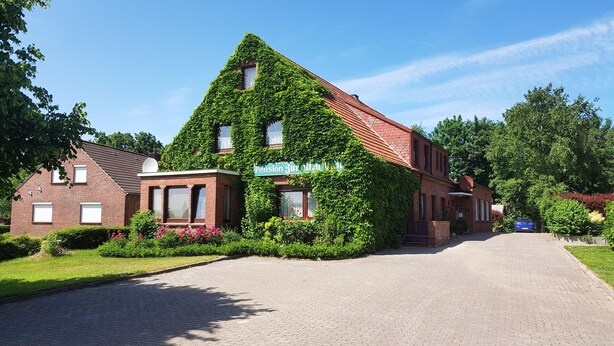 A building with a sloped roof covered in green ivy, a sign reading "Pension Zur alten Wiese," and a paved driveway leading to the entrance. - Gästehaus Zur Alten Post Wohnung Ost (Dornum)