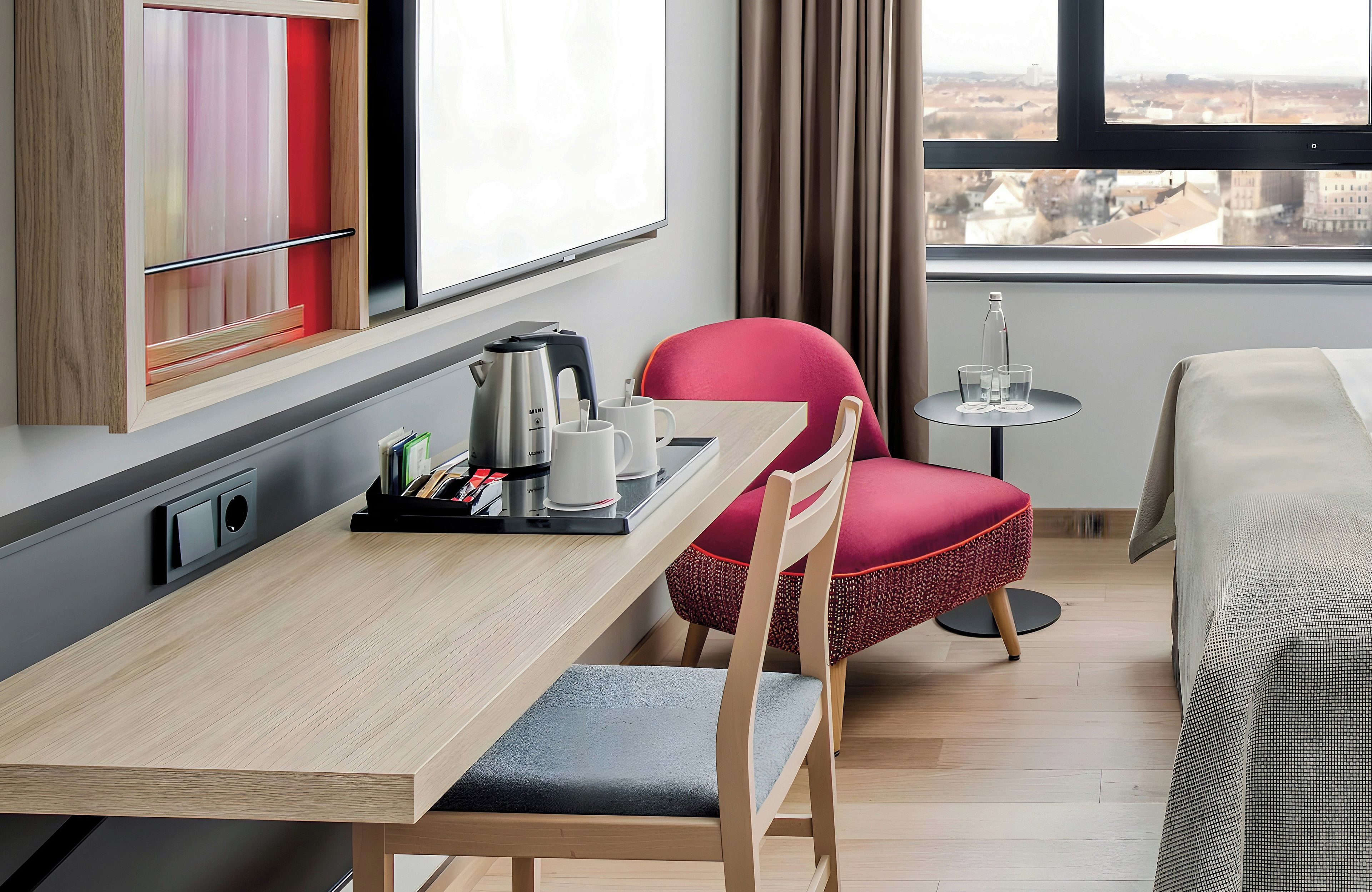 Een hotelkamer met een houten tafel, een rode stoel, een bed en uitzicht op de stad door het raam.