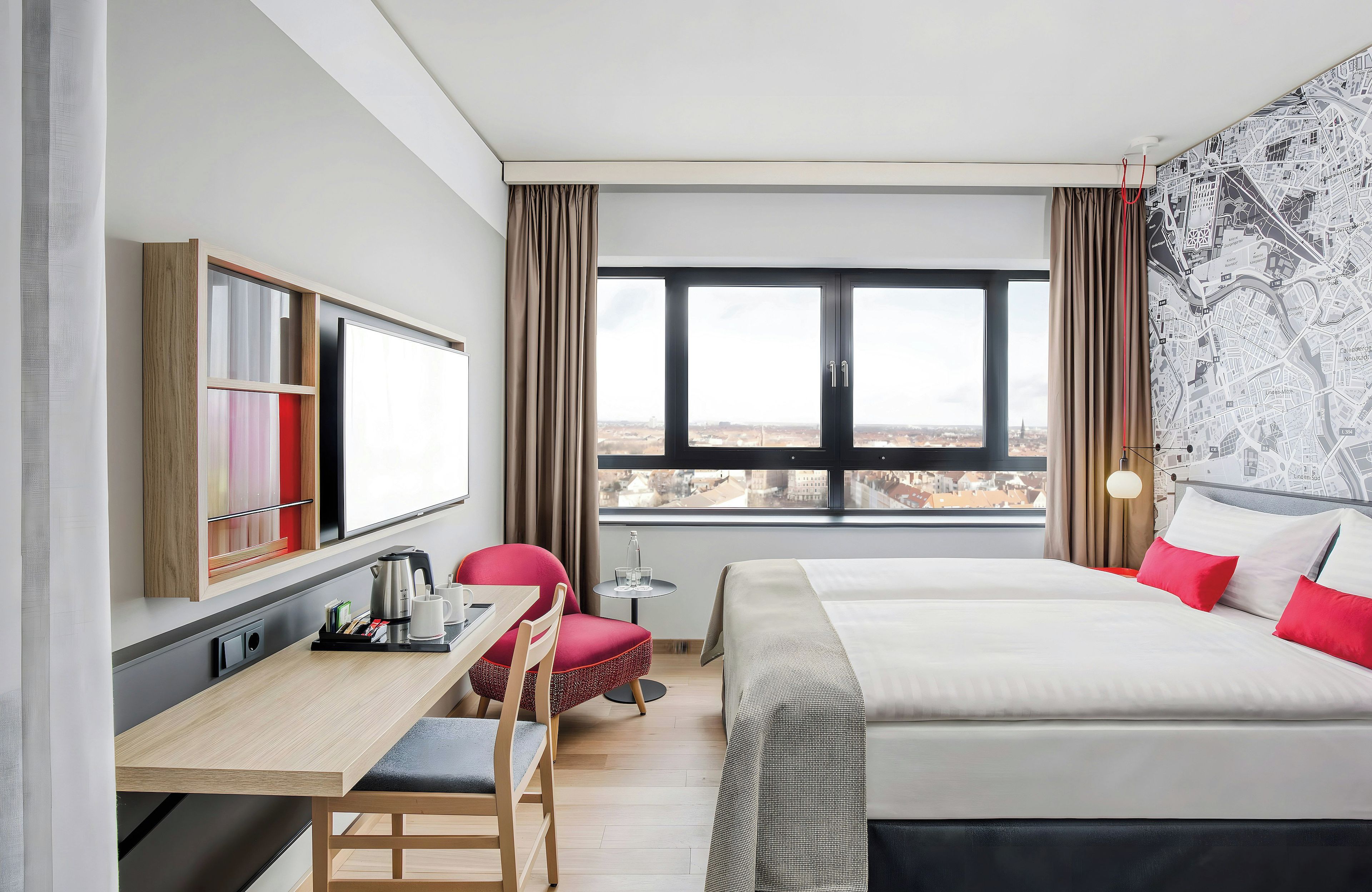 Ein Hotelzimmer mit einem Bett, einem Schreibtisch mit Wasserkocher, einem Sessel, einem kleinen Tisch mit Gläsern und einem Fenster mit Blick auf die Stadt.