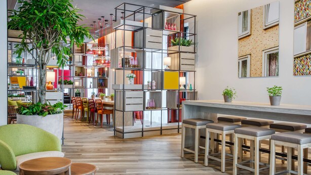 Un interior moderno con una zona de estar central, una barra con taburetes y una estantería escalonada con diversos objetos.