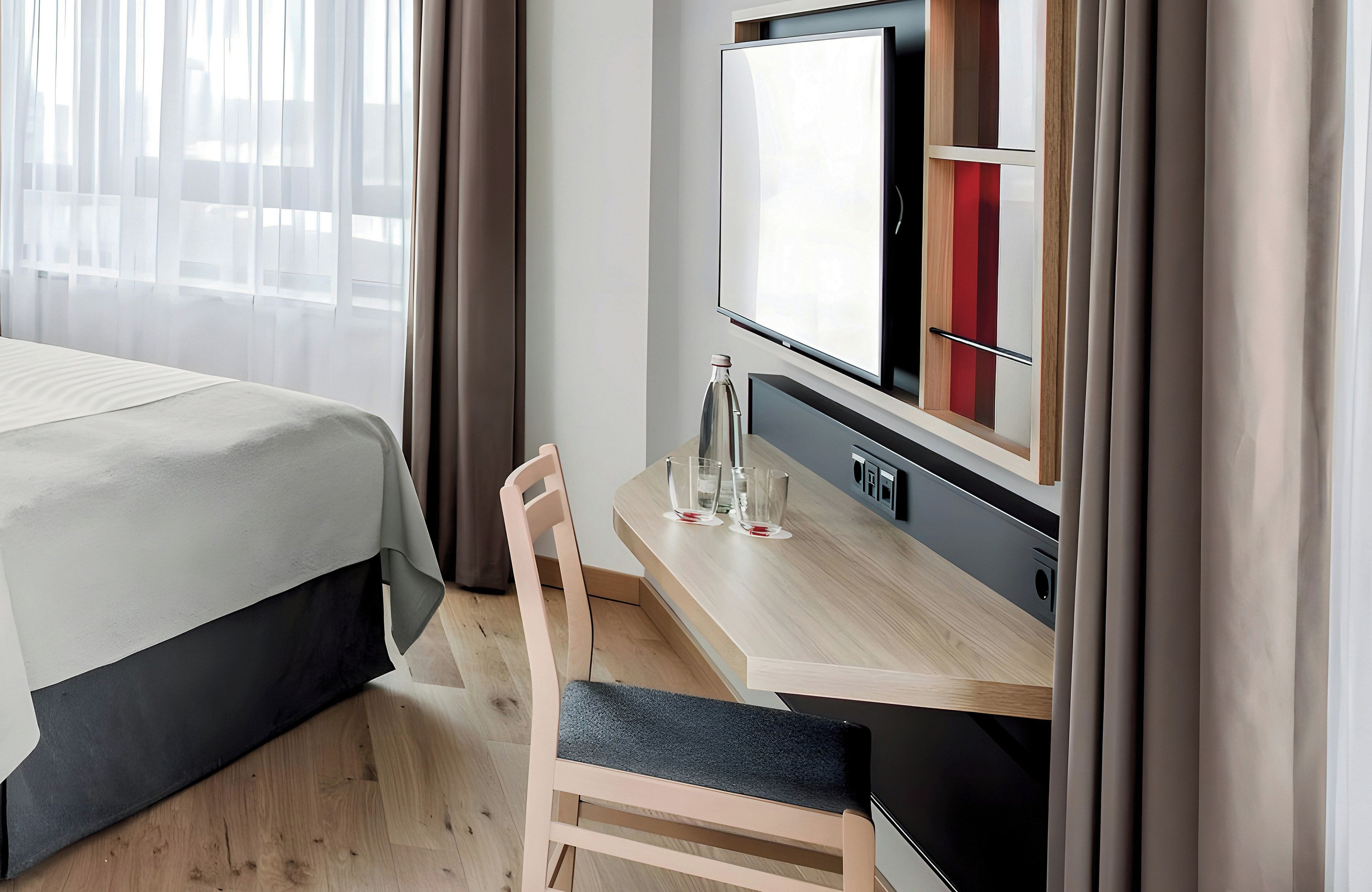 Hotellihuone, jossa on sänky, työpöytä tuolilla, televisio ja ikkuna, jossa on verhot.