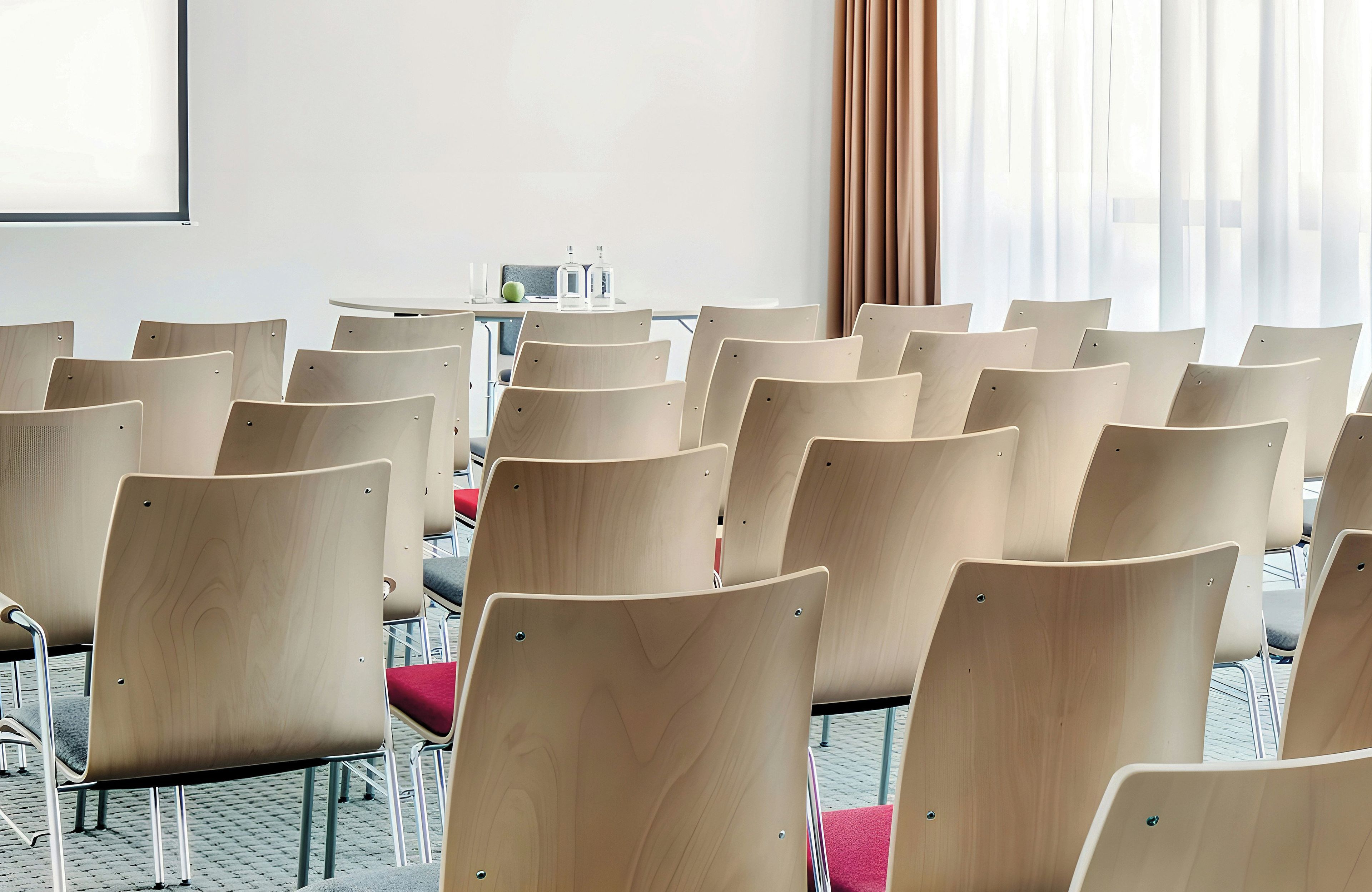 Ein Konferenzraum mit Reihen von Stühlen, einem Tisch mit einem Laptop und einem Fenster mit Vorhängen.