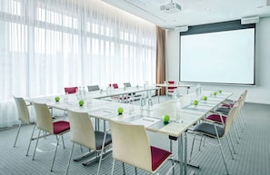 A conference room with a long table, chairs, a projector screen, and a large window with curtains. - Leonardo Hotel Hannover City Center (Hannover)