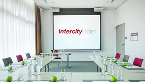Una sala de conferencias en el Hotel Intercity, con una pantalla grande que muestra el nombre del hotel, una mesa larga con botellas de agua y manzanas, y varias sillas alrededor.