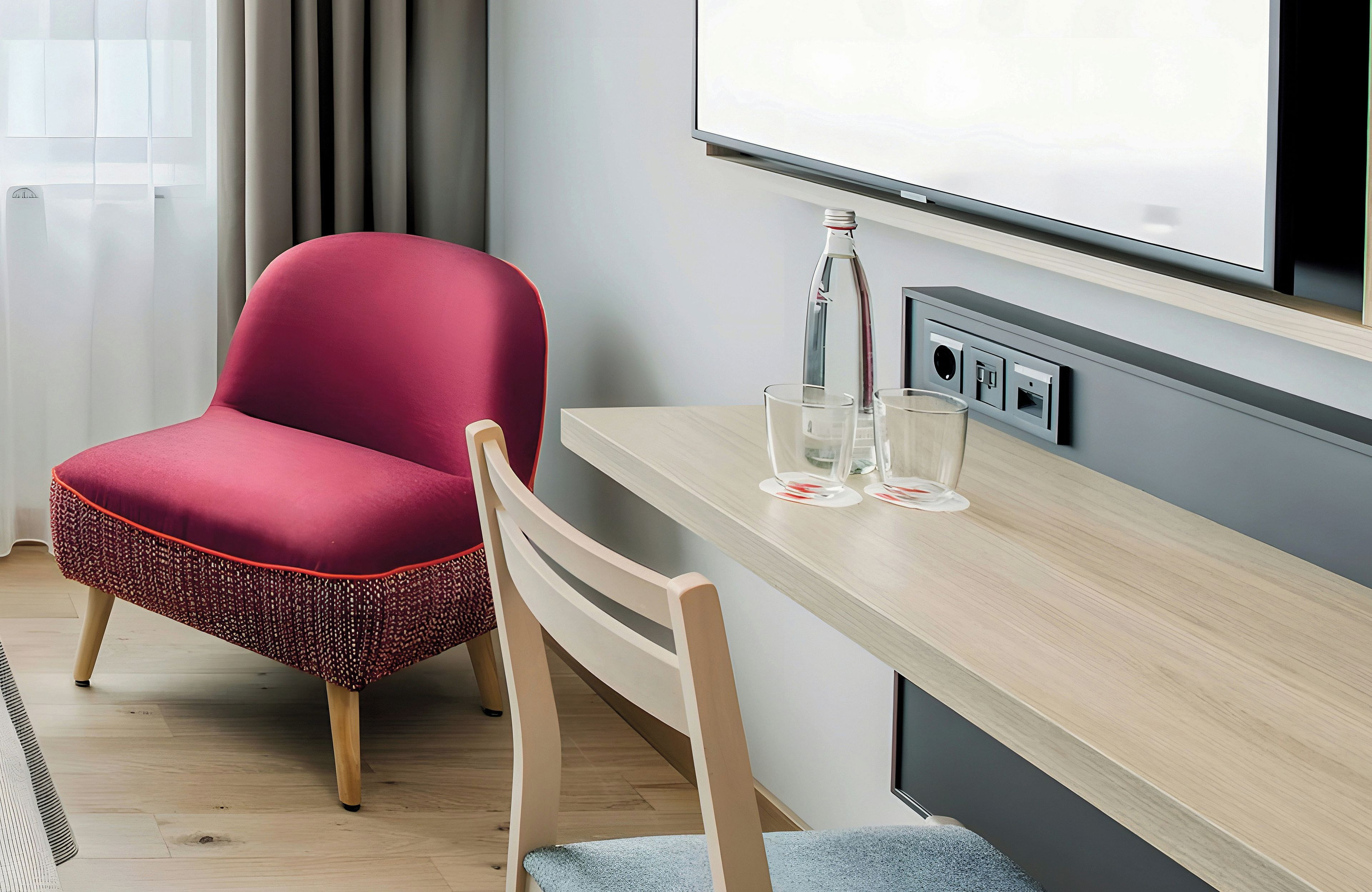 A modern hotel room with a wooden desk, a red upholstered chair, a glass table, and a flat-screen TV mounted on the wall.