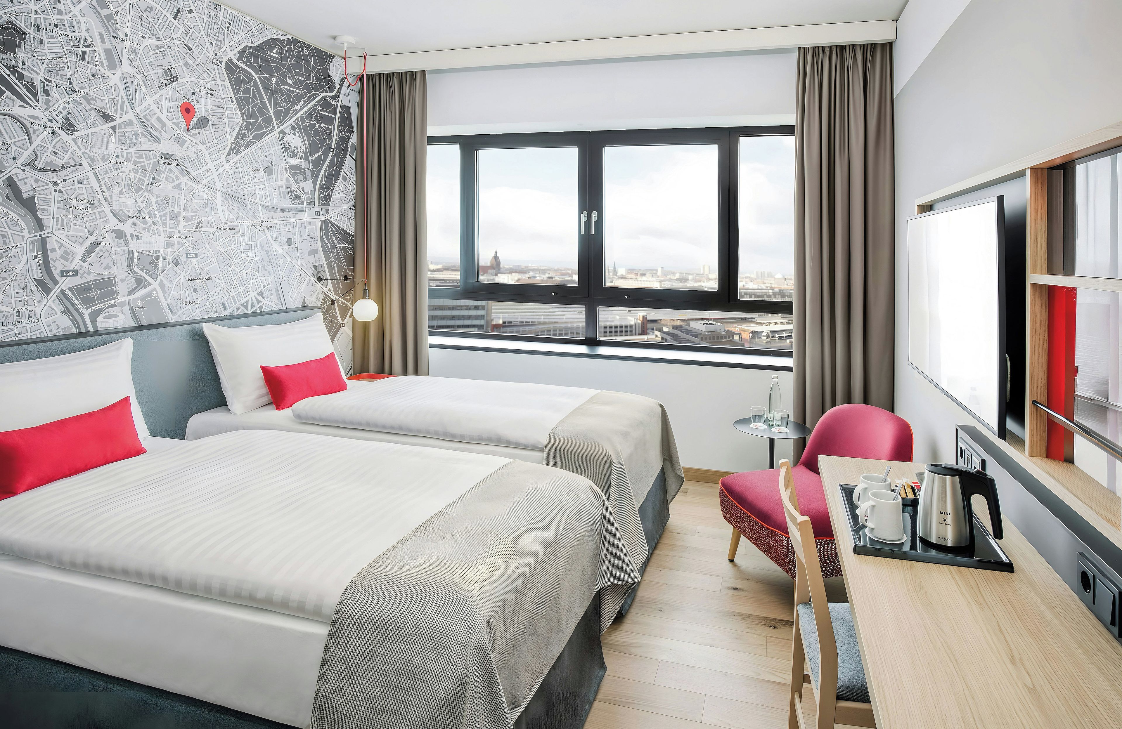 A hotel room with two beds, a desk with a chair, a view of the city through a large window, and a map on the wall.