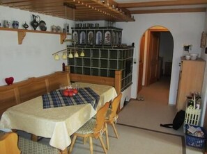 A dining area with a table covered in a checkered tablecloth, wooden chairs, and a bowl of apples. There is a green tiled cabinet with a mirror and a shelf above it. - Aalener Haus Wohnung 3 (Oberstdorf)