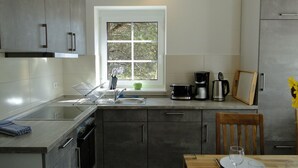 A modern kitchen with grey cabinets, a built-in oven, a sink, and a window with a view of trees. There is a dining table with a vase of sunflowers. - Bispinger Heidezauber - Haus Hartriegel (Bispingen)