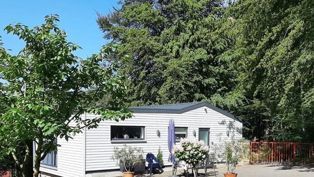 Una pequeña casa móvil de un piso con techo plano, un porche delantero y un pequeño jardín con plantas en macetas.