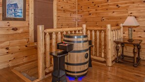 Interior de una cabaña de madera con un barril de madera, una mesita con una lámpara y un cuadro en la pared.