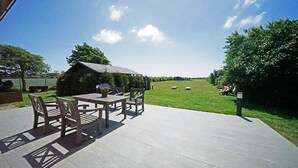 Un patio al aire libre con una mesa y sillas, un área de estar techada, y vistas a un campo verde y árboles.