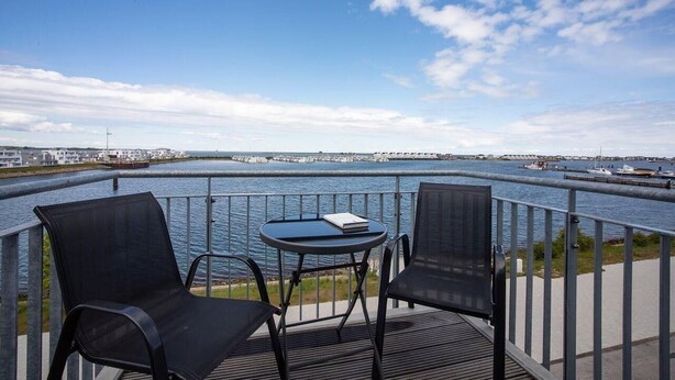 A balcony with two chairs and a small table overlooking a body of water with boats and a distant shoreline. - Penthouse-hafenwohnung Reeders Topp (Kappeln)