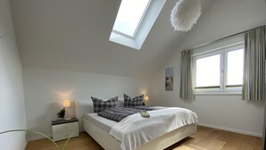 A modern bedroom with a skylight, a bed with a white comforter and checkered pillows, a bedside table with a lamp, and a window with curtains. - Penthouse-hafenwohnung Reeders Topp (Kappeln)