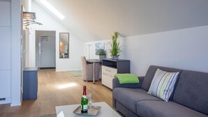 A modern living room with a grey sofa, a small table with a bottle and glass, a kitchenette, and a dining area with a patterned chair. - Penthouse-hafenwohnung Reeders Topp (Kappeln)
