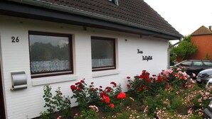 A white building with the number 26 and the name "Haus Kalliglick" on the facade. There is a garden with pink and red flowers in front of the building. - Haus Halligblick, Ferienwohnung Pellworm (Dagebuell)