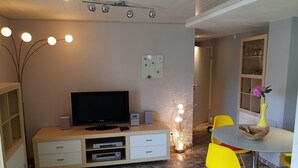 Un saló modern amb televisió de pantalla plana, un moble baix, una llum decorativa i una zona de menjador amb una taula i cadires grogues.