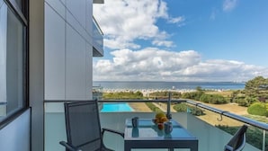 En balkon med et glastelt bord, to stole og udsigt til havet og fjerne bygninger.