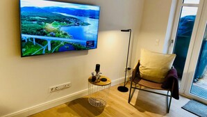 A room with a mounted TV displaying a scenic landscape, a standing lamp, a wicker chair, and a small table with decorative items. - Ankerplatz-sylt Haus Mateika - Wohnen Sie Exklusiv im Herzen von Westerland (Sylt)