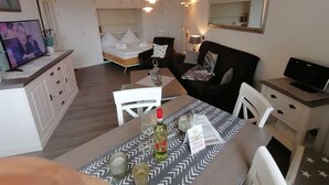 A modern living room with a dining area, a bed with white linens, a TV, and a mirror on the wall. - Nordmarkhof (Sylt)