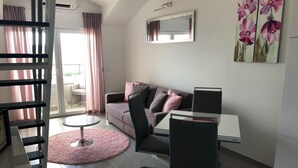 A modern living room with a sofa, a dining table, and a chair. There is a staircase on the left, a window with curtains, and a painting on the wall. - Studioapartment "carpe Diem" (Kastela)