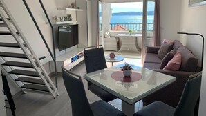 A modern living room with a glass table, a sofa, and a TV on a white cabinet. A balcony with a view of the sea is visible through an open door. - Studioapartment "carpe Diem" (Kastela)