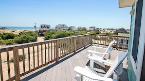 A wooden deck with white chairs, overlooking a beach with houses in the distance.