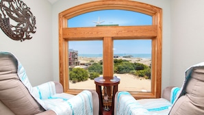 A room with a large window overlooking a beach, two cushioned chairs, a decorative wall piece, and a small wooden table.