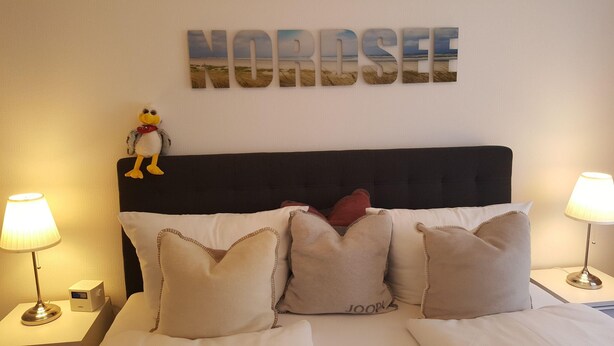 A bed with a dark headboard, multiple pillows, a decorative bird, and a 'NORDSEE' sign on the wall. - App. Syltliebe, Haus Ankerlicht, Whg. 70 (Sylt)