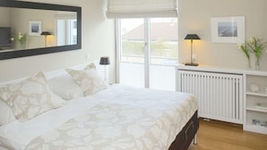 A bedroom with a large bed, a bedside table with a lamp, a radiator, and a window with a view. - Victoriaresidenz App. 12, Ekki' s Suite (Sylt)