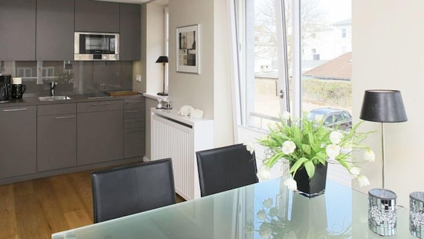 A modern kitchen with grey cabinets, a built-in microwave, and a dining area with a glass table and chairs. There is a window with a view of the outside. - Victoriaresidenz App. 12, Ekki' s Suite (Sylt)
