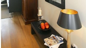 A modern living room with a wooden floor, a black sofa, a wooden side table with a bowl of fruit and magazines, a black and gold lamp, and a large mirror on the wall. - Victoriaresidenz App. 12, Ekki' s Suite (Sylt)