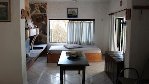 A room with a bed, a table, a chair, a window with curtains, a stone wall, and a painting on the wall.