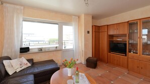 A living room with a sofa, a coffee table with a vase of flowers, a television, and wooden cabinets. - Wohnung Lee (Sylt)
