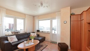 A living room with a brown leather sofa, a wooden coffee table, and a view of the city through large windows. - Wohnung Lee (Sylt)